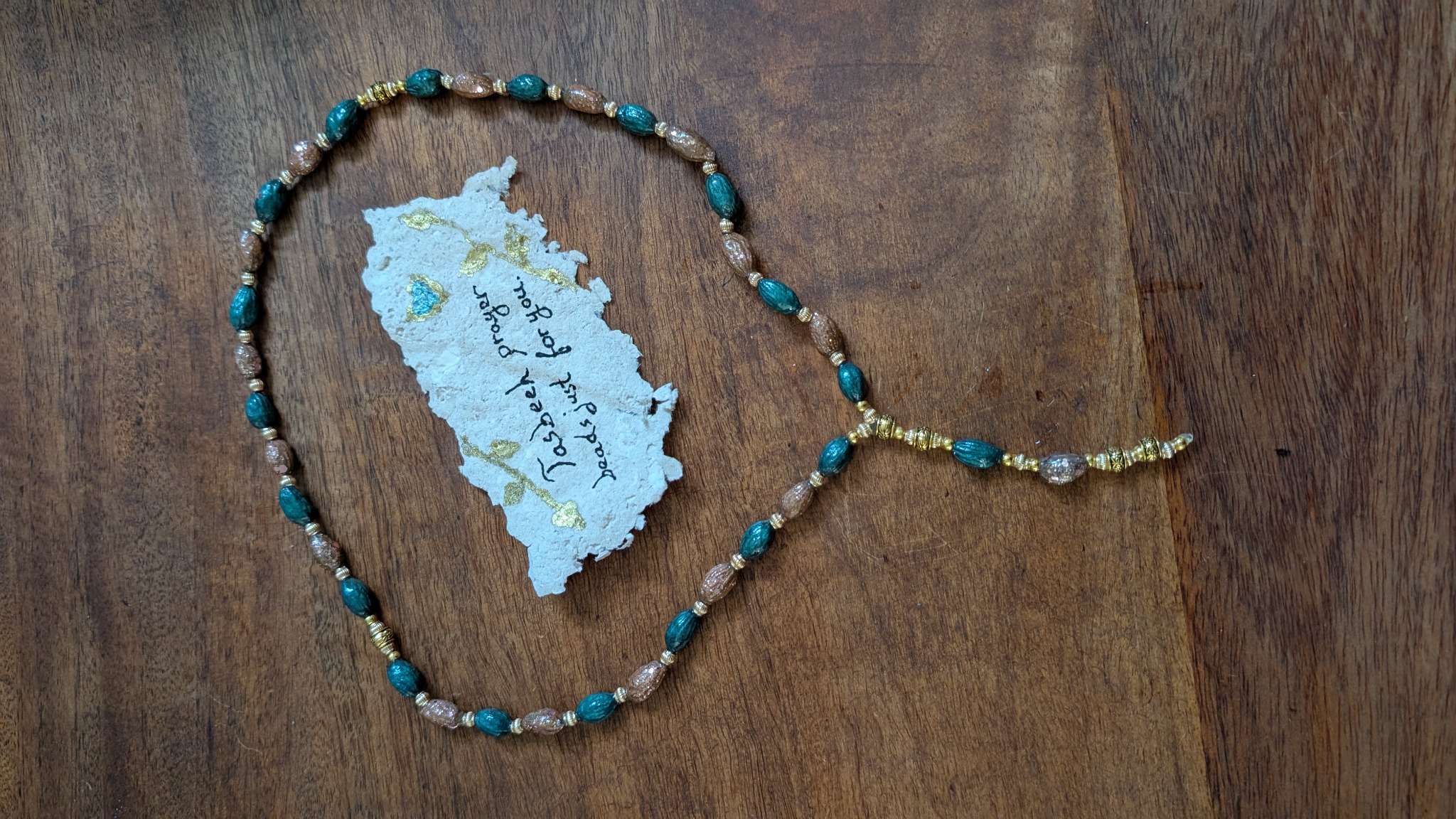 Handmade tasbeeh prayer beads using painted and sealed olive pits in teal and warm bronze tones, with golden spacer beads and a handwritten note