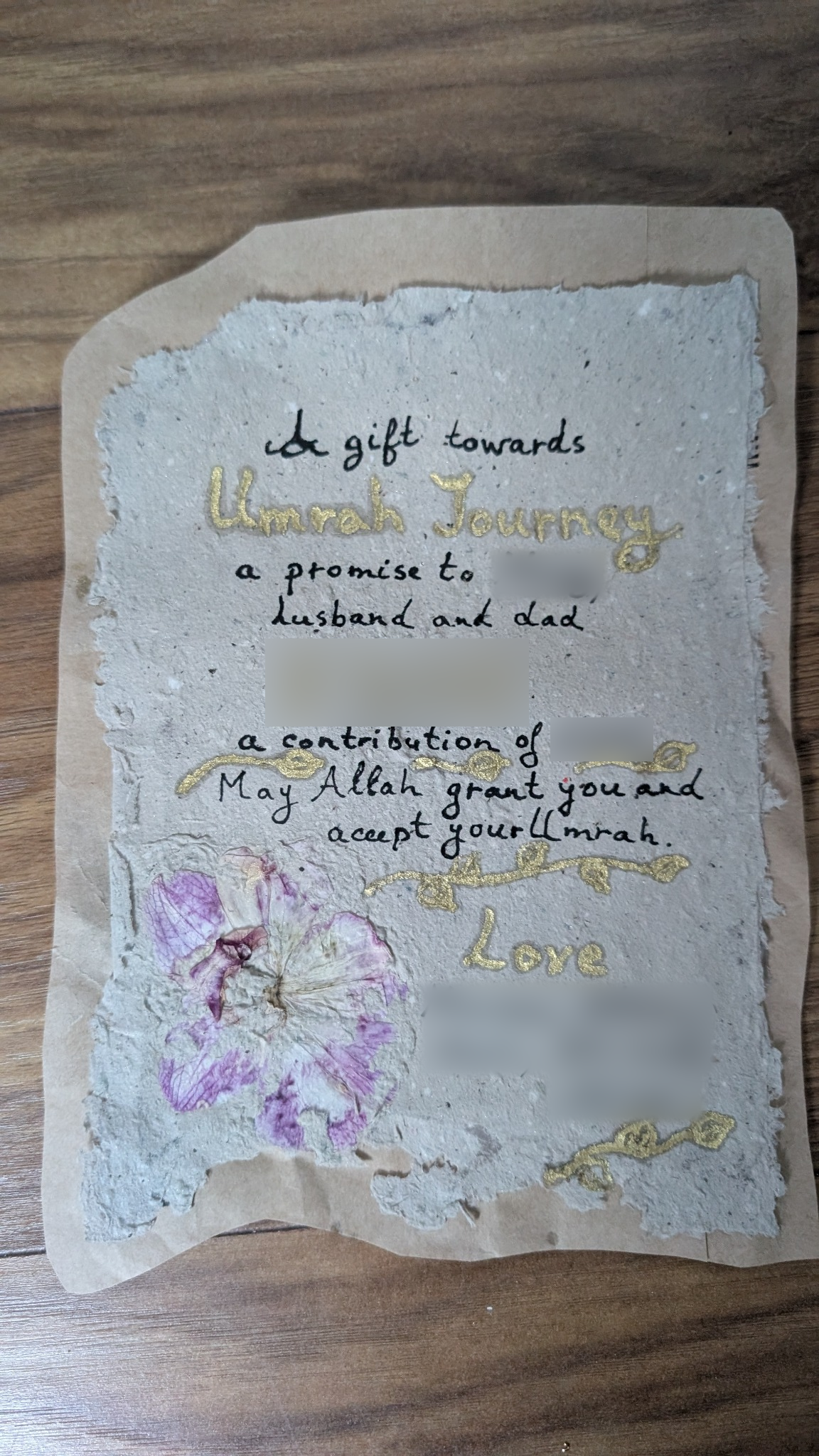 Original handmade promise note with a pressed birthday orchid, gold details and upcycled envelope backing; family names and gift value blurred